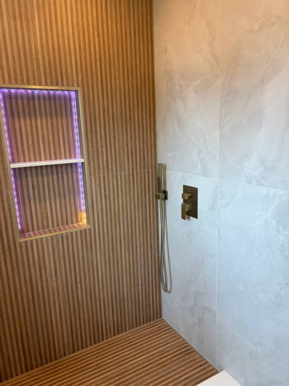 Wide angle of wood slat wall with LED niche, marble wall, and brass shower valve